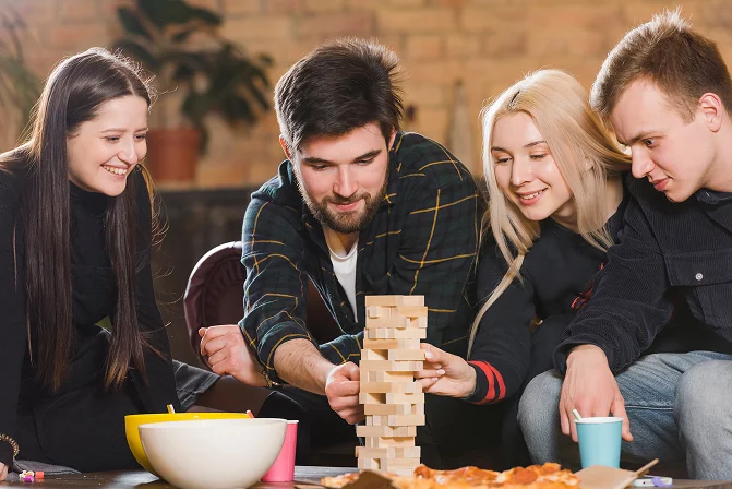 Skupina čtyř mladých přátel se usmívá a soustředěně hraje stolní hru Jenga u stolu s jídlem a pitím.