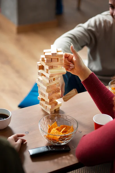Pohled shora na dřevěnou věž Jenga, ze které padá blok, na stole s miskou chipsů a dálkovým ovladačem.